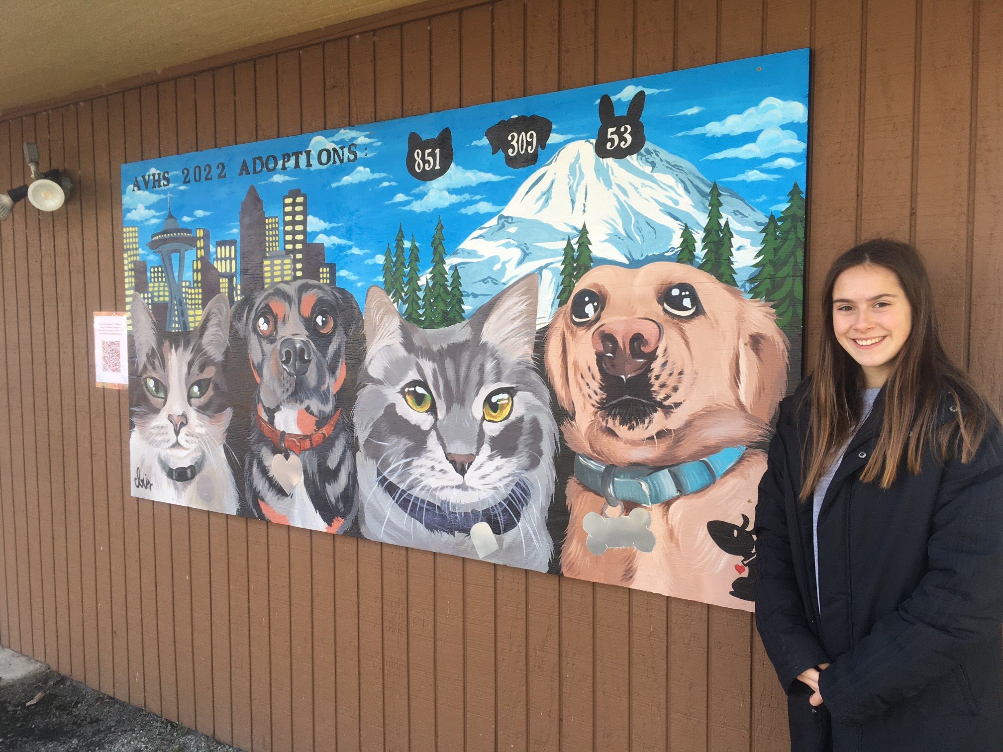 A mural featuring painted images of cats and dogs on the side of a building, labeled AVHS 2022 Adoptions, with a woman standing to the right and smiling.